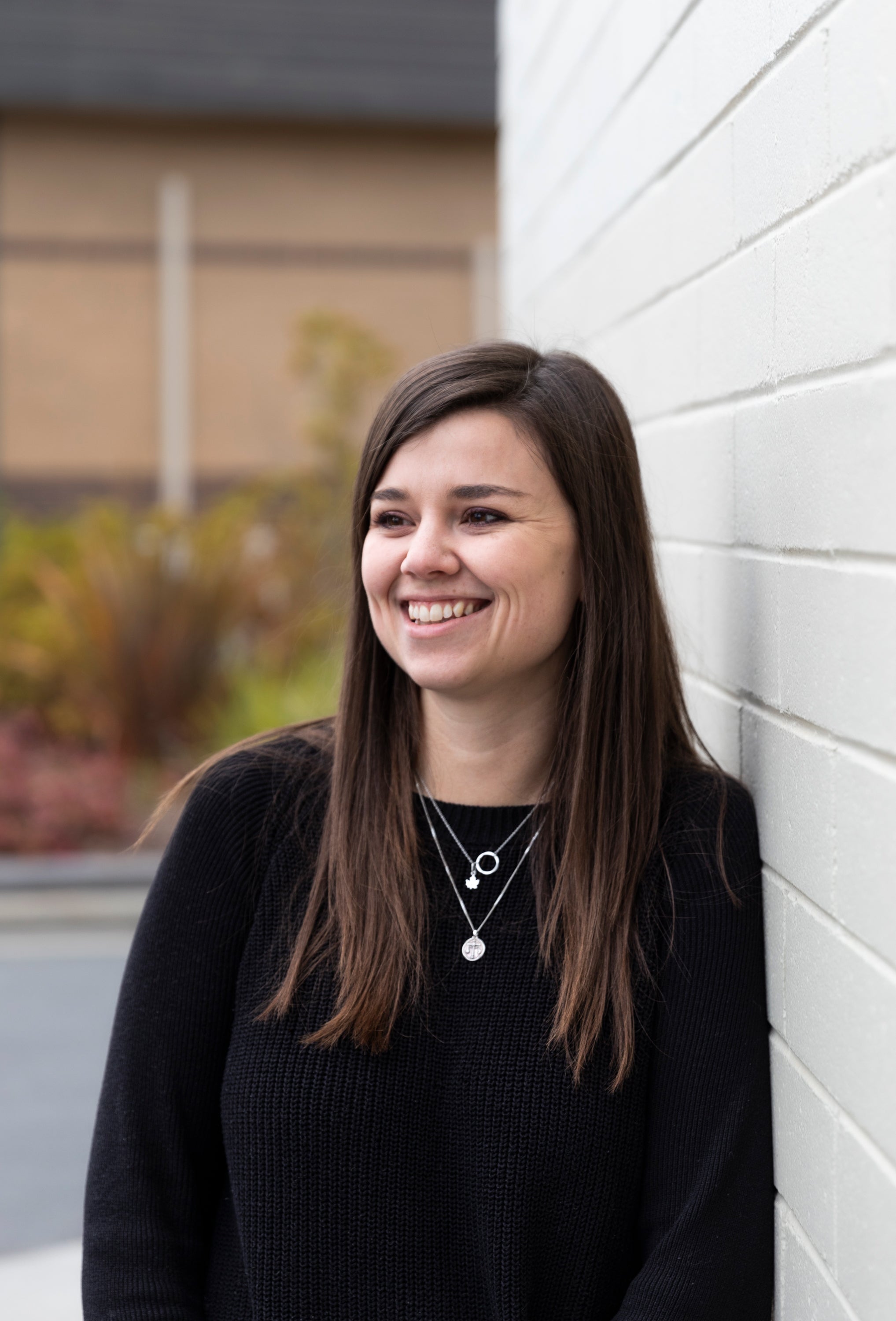 woman smiling with long dark brown hair leans against a white wall in a black top