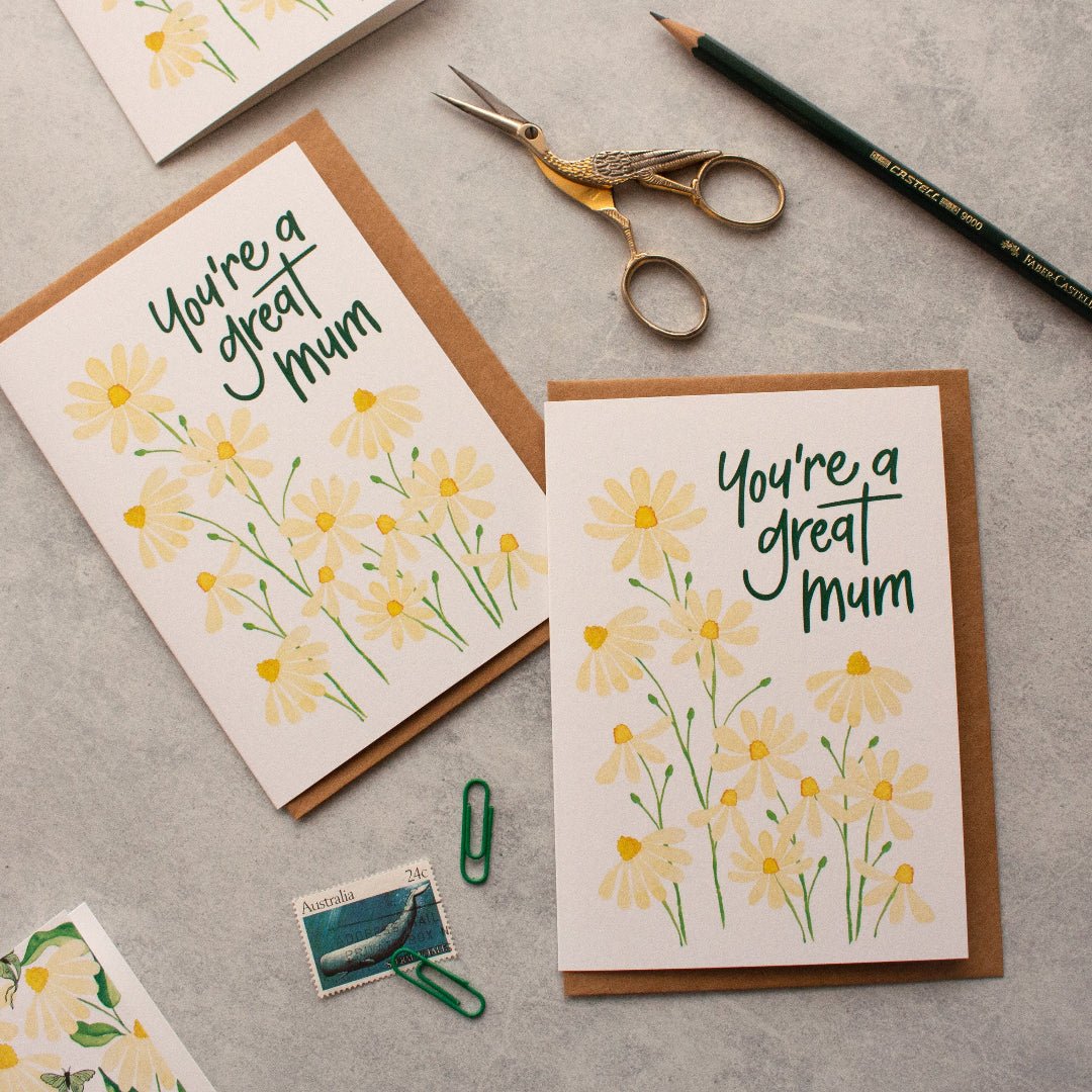 Two mum greeting cards with daisy designs and text on a grey surface with stationery items.