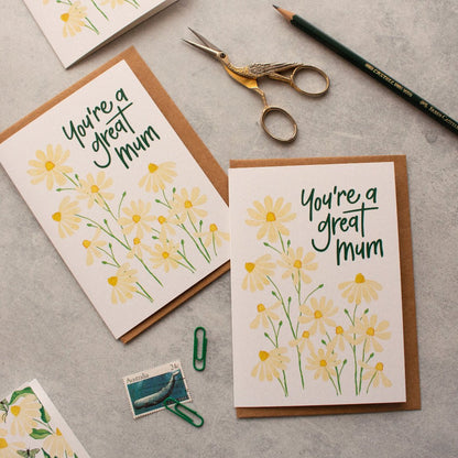 Two mum greeting cards with daisy designs and text on a grey surface with stationery items.