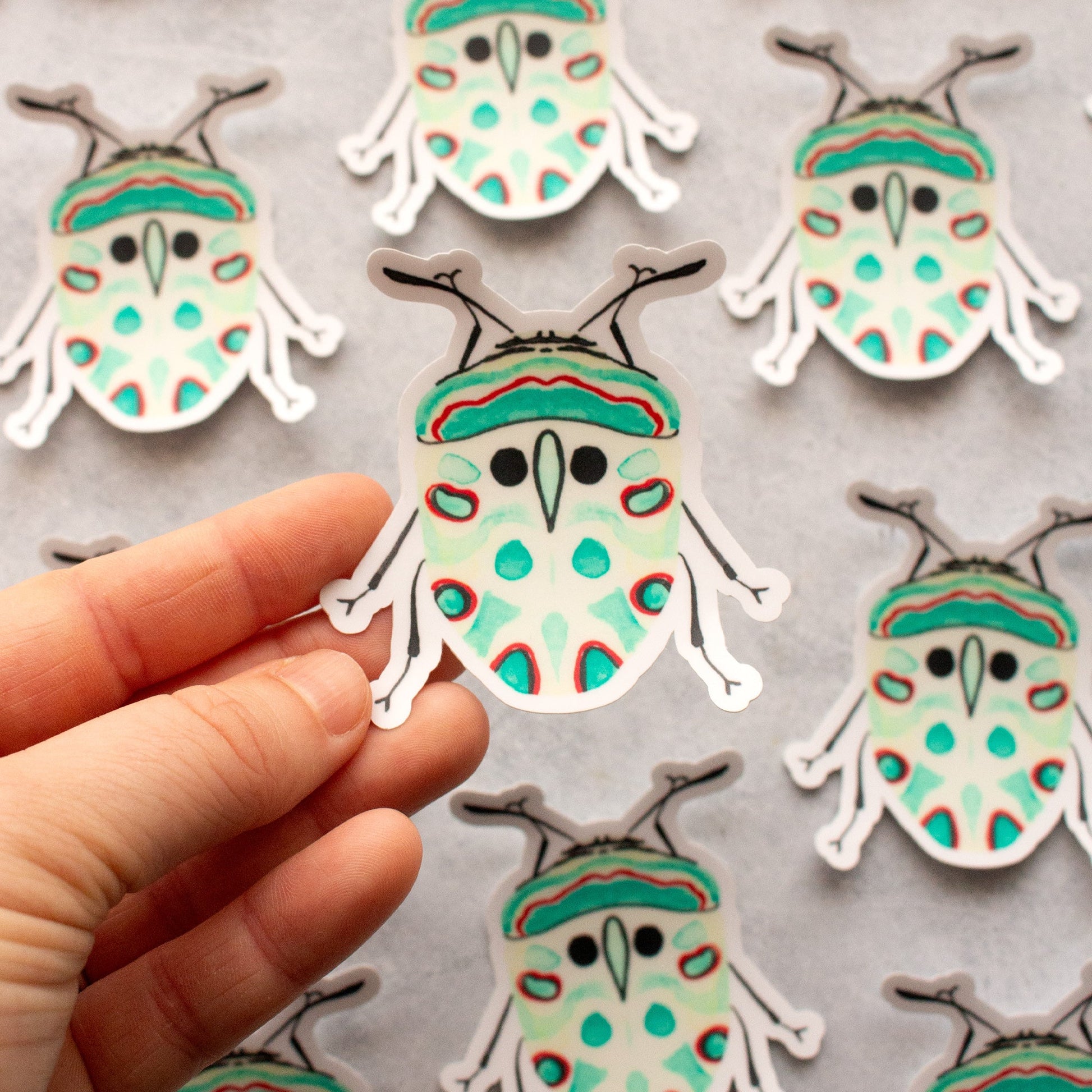 Hand holding a colourful beetle-shaped sticker with more stickers in the background on a grey surface.