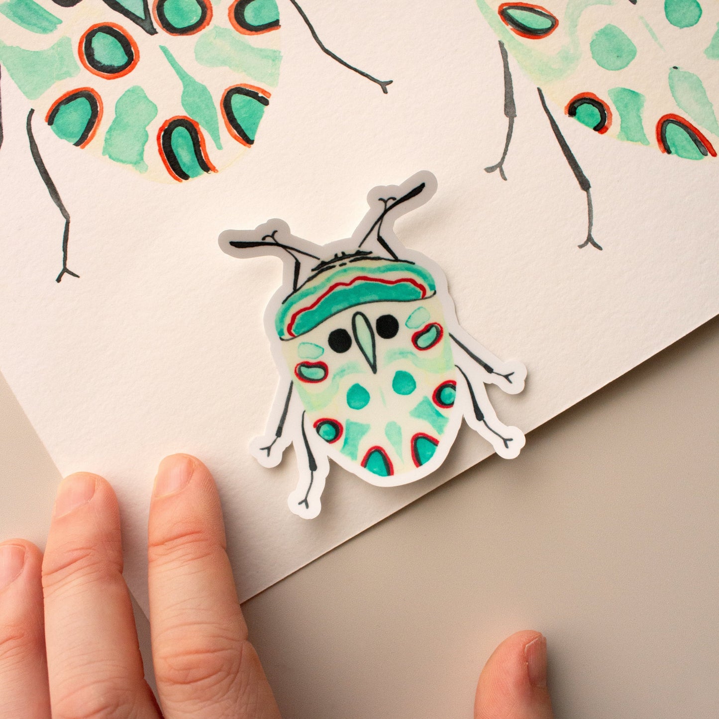 Colourful beetle sticker held by a hand with a matching design on paper in the background.