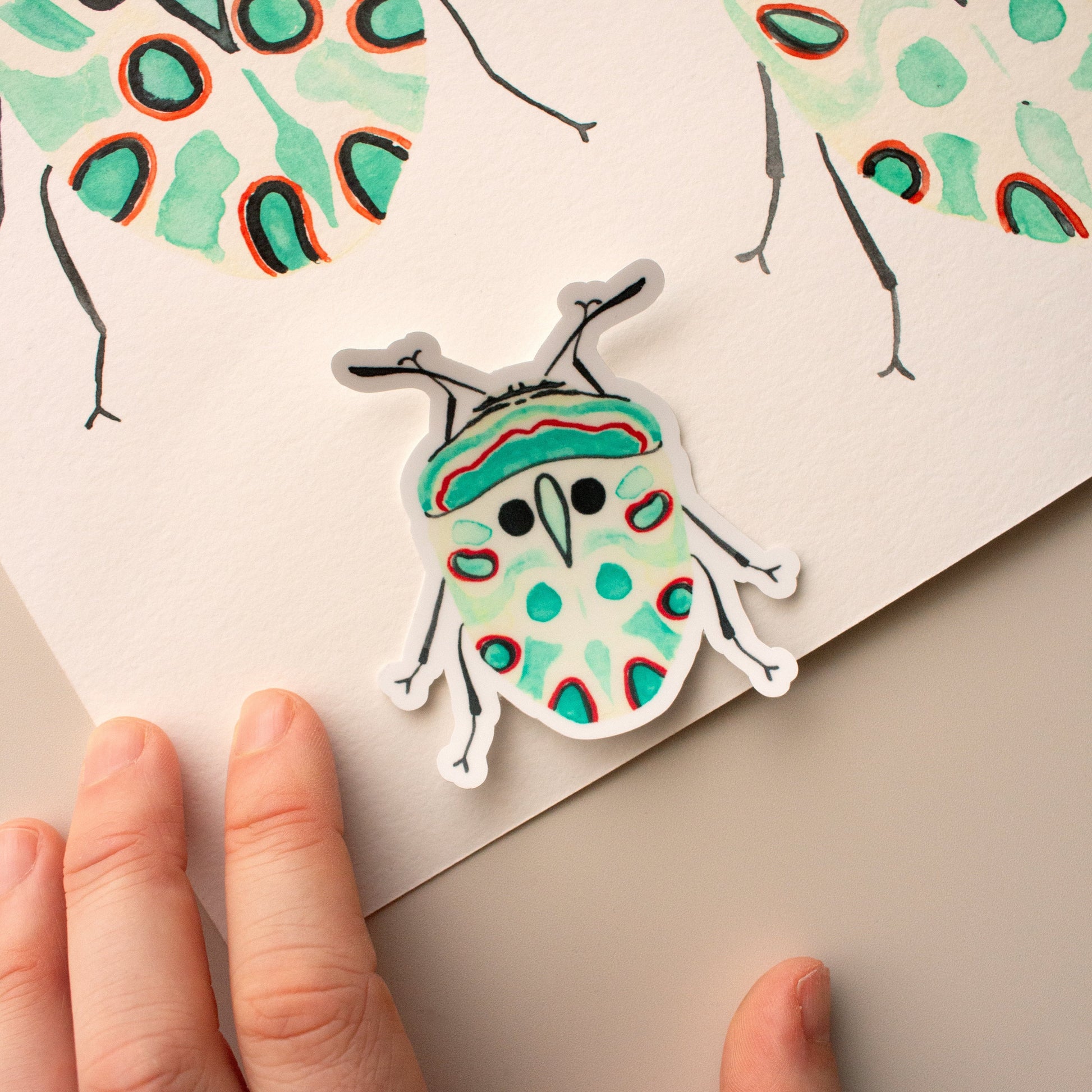 Colourful beetle sticker held by a hand with a matching design on paper in the background.