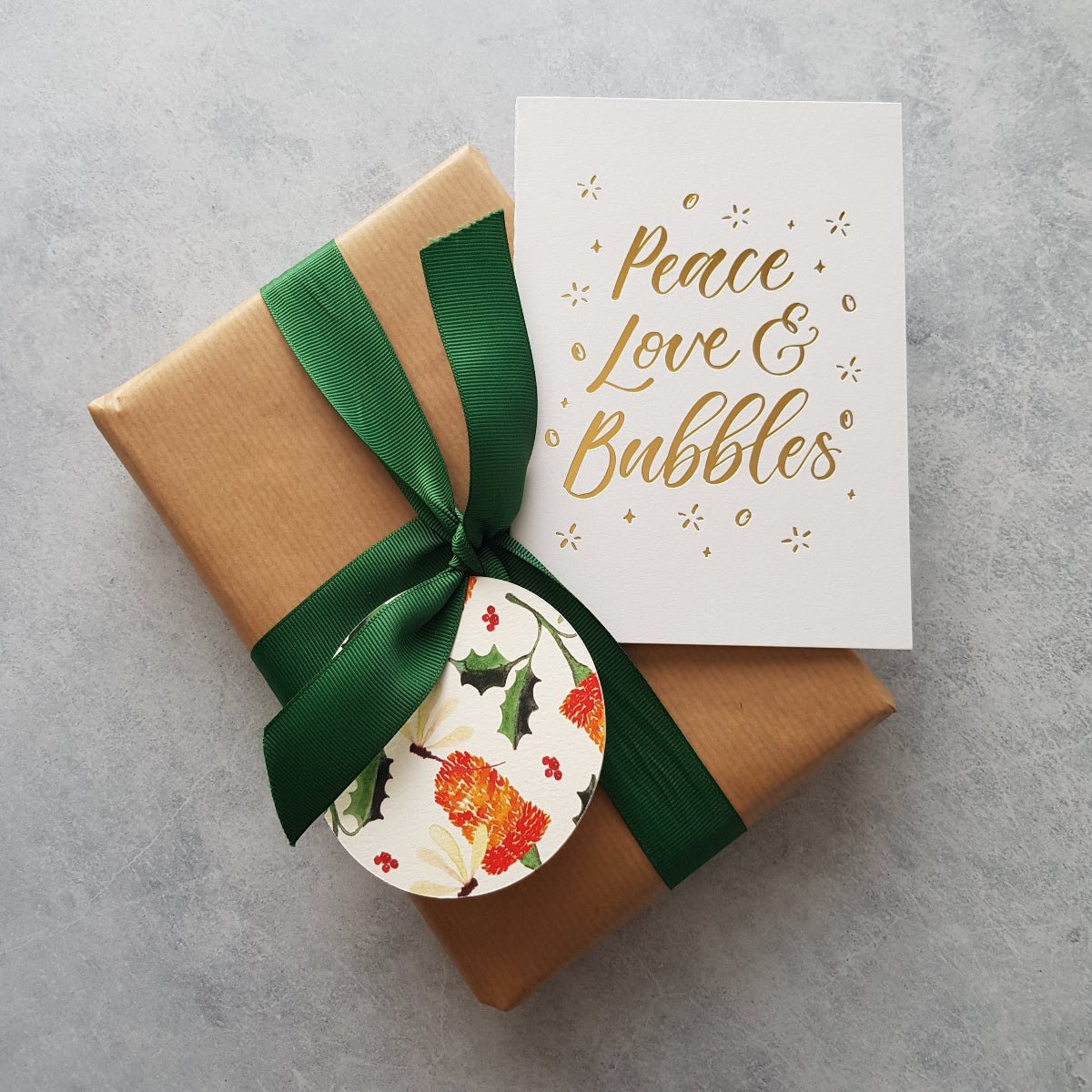 A christmas card with the words 'peace, love and bubbles' in gold foil on a present wrapped in brown with a green ribbon