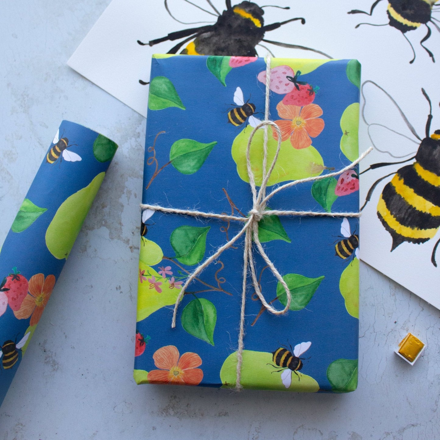 Present wrapped in pears, flowers, strawberries and bees tied in string. To the left of the present is a single roll of wrapping paper. The present sits on top of a large sheet of paper with hand painted bees. 