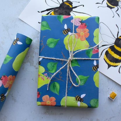 Present wrapped in pears, flowers, strawberries and bees tied in string. To the left of the present is a single roll of wrapping paper. The present sits on top of a large sheet of paper with hand painted bees. 