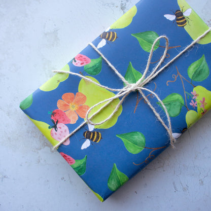 Wrapped present sits on top of a stone background. The wrapping paper design has a blue background with repeat pattern of green pears, orange poppy flowers, pink strawberries, green leaves and yellow bees. The present is tied in white string in the shape of a bow.
