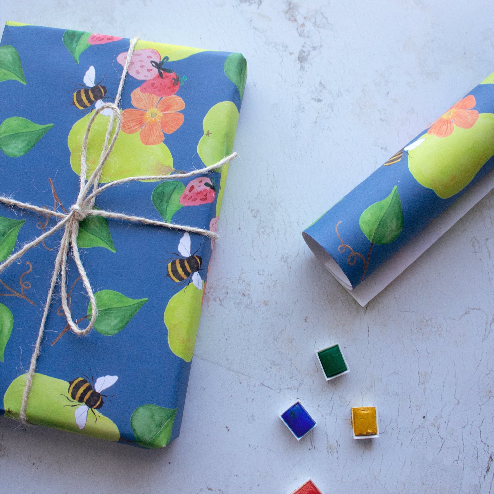 Gift on the left hand side wrapped in repeat pattern of pears, bees, strawberries and poppy flowers. The gift has a tied bow around the present and is surrounded by a roll of wrapping paper and little paint trays