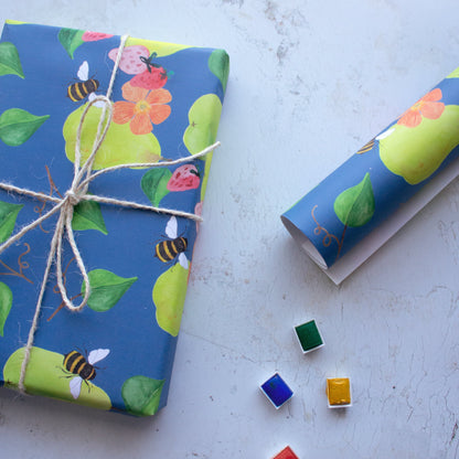Gift on the left hand side wrapped in repeat pattern of pears, bees, strawberries and poppy flowers. The gift has a tied bow around the present and is surrounded by a roll of wrapping paper and little paint trays