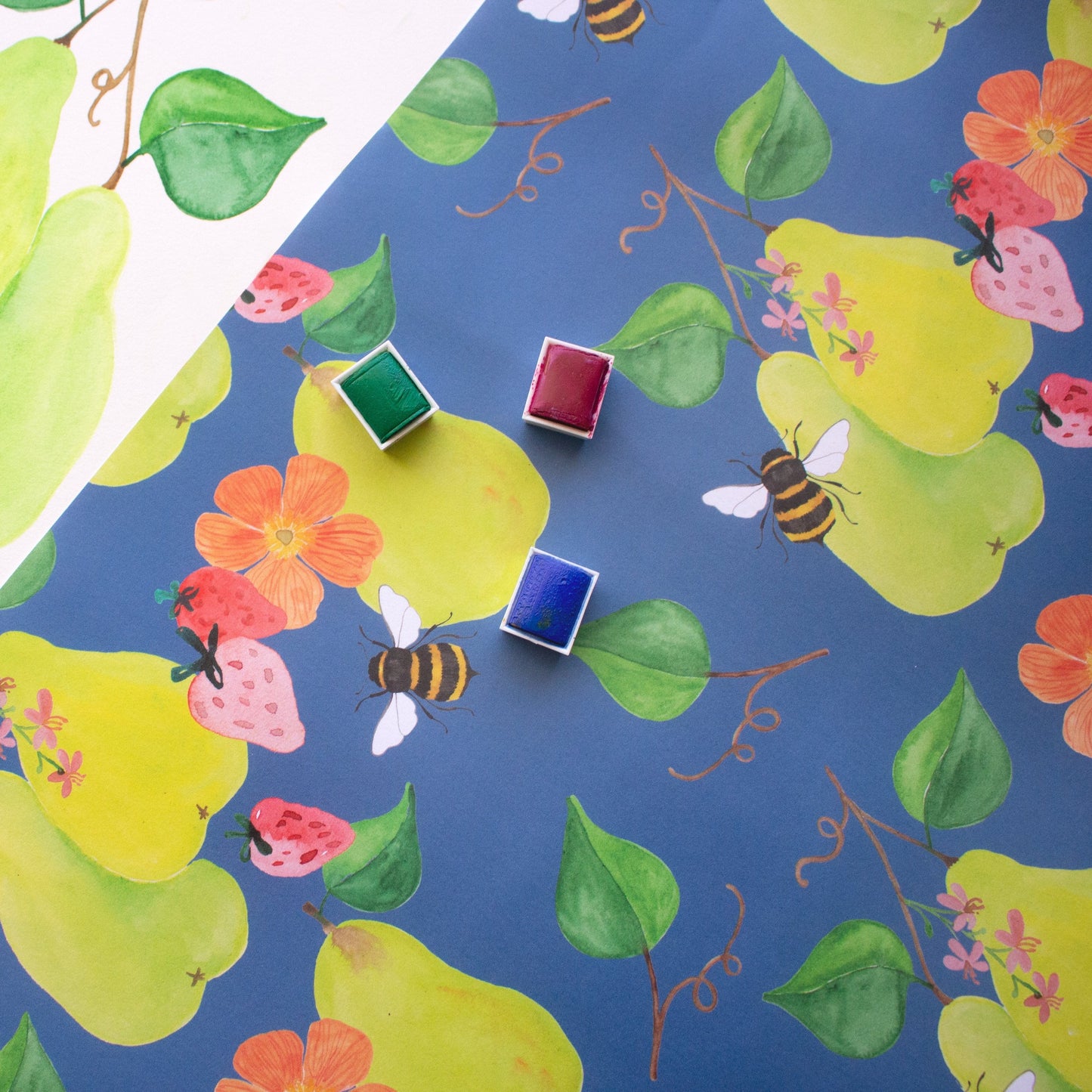 blue patterned print with green floral pears and pink strawberries. Three watercolour trays in green, red and blue sit on top of the paper. 