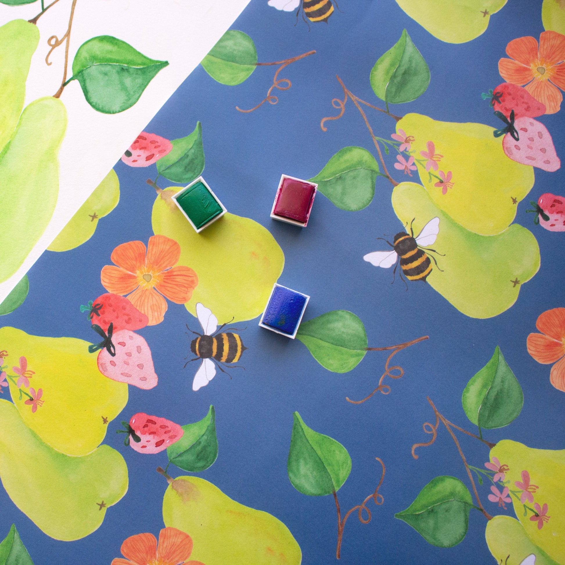 blue patterned print with green floral pears and pink strawberries. Three watercolour trays in green, red and blue sit on top of the paper. 