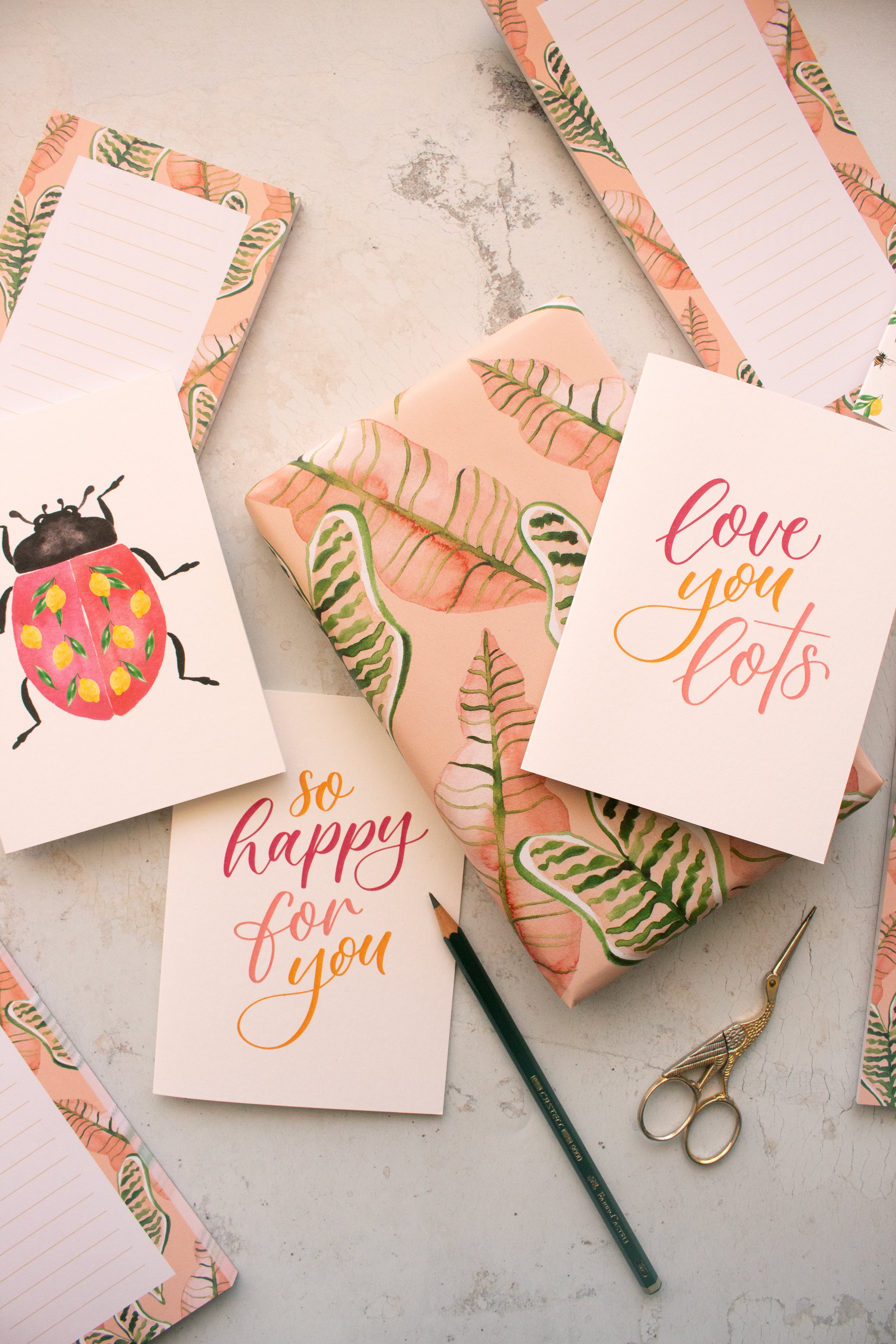 Pink palm leaves notepads and gift wrap surrounded by stationery, scissors, a pencil and greeting cards with pink and yellow scripts and a giant lady bug.