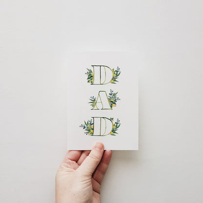 Hand holding greeting card DAD' surrounded by leaf designs on a white background