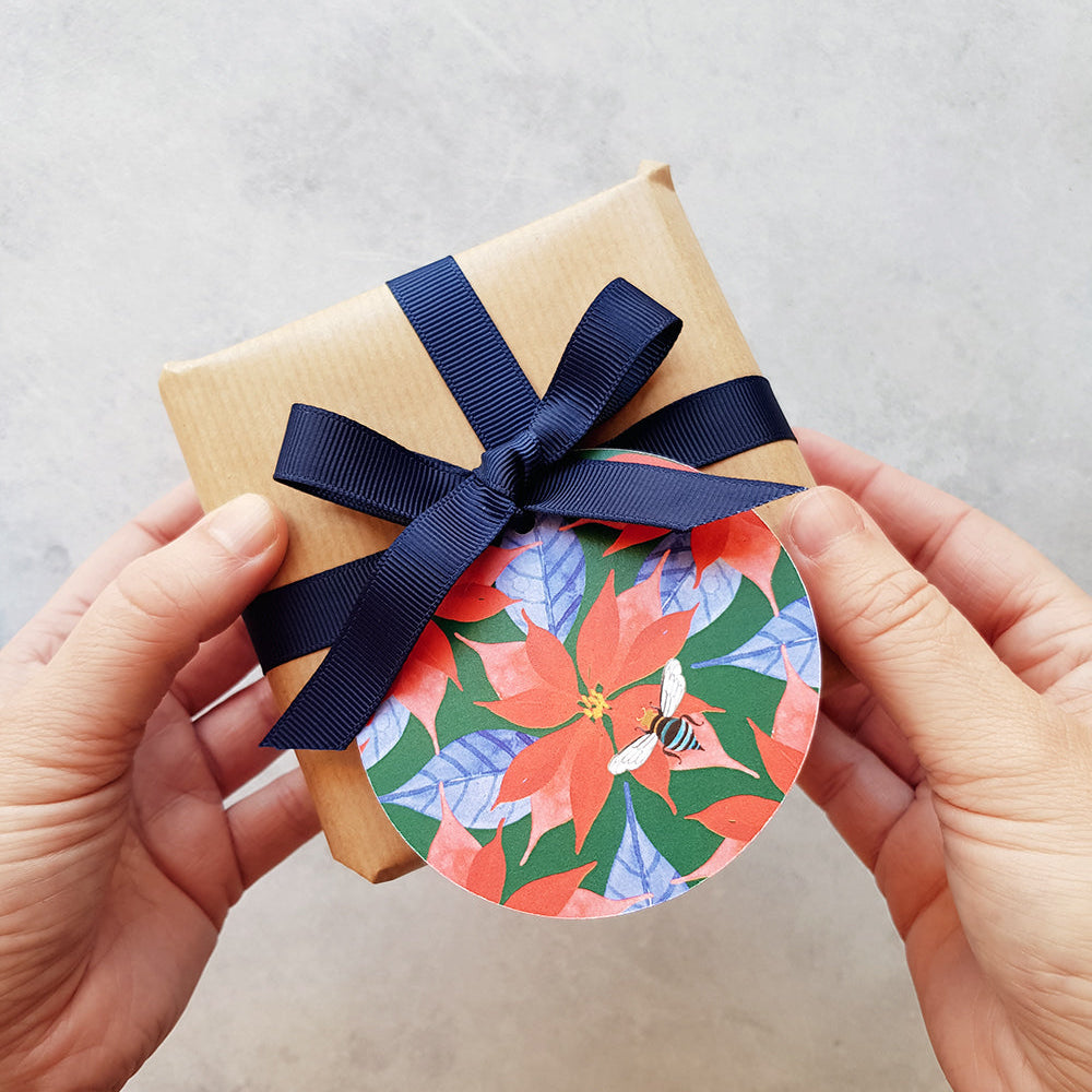 A person's hands holding a small kraft paper gift box tied with a navy blue ribbon, featuring a colorful floral paper tag with red, green, and blue poinsettia-style flowers.