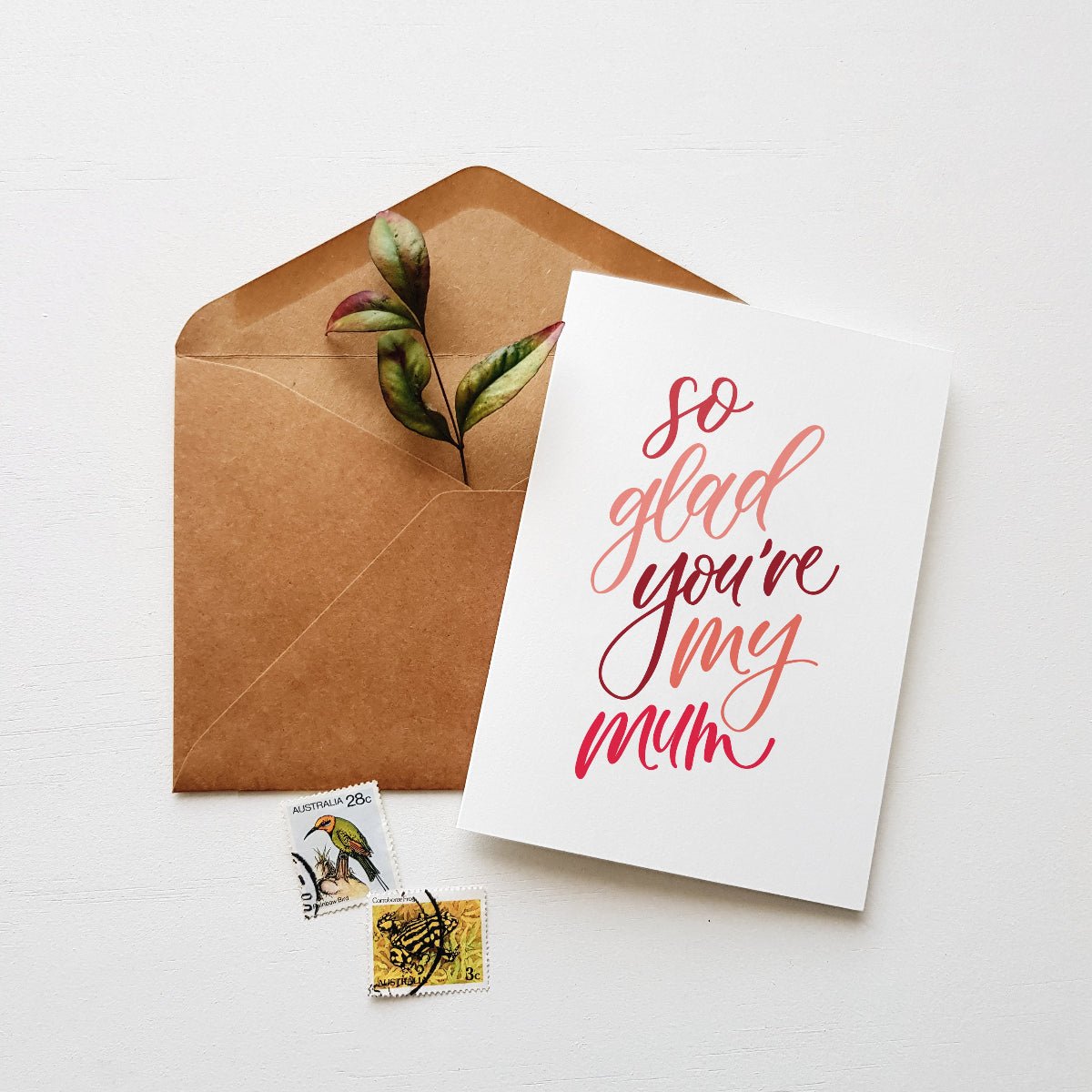 Greeting card with 'so glad you're my mum' text, brown envelope, and stamps on a white background