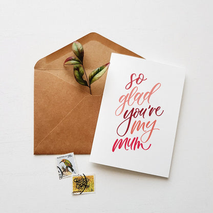 Greeting card with 'so glad you're my mum' text, brown envelope, and stamps on a white background