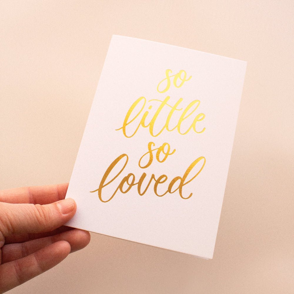 Hand holding a white card with 'so little so loved' in gold text on a beige background