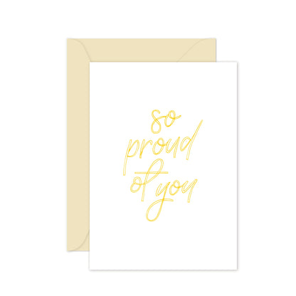 A white greeting card with yellow hand lettering that reads 'so proud of you', accompanied by a cream envelope.