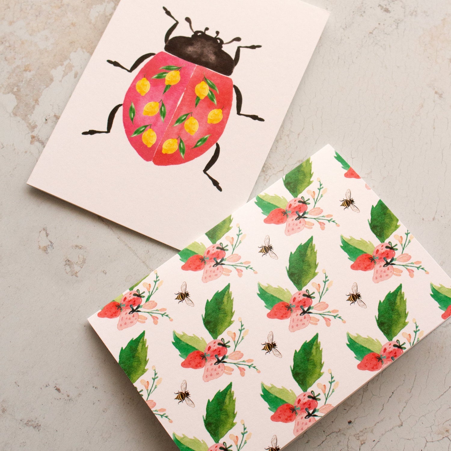Two greeting cards on a textured surface, one with a ladybug design and the other with floral and insect pattern.