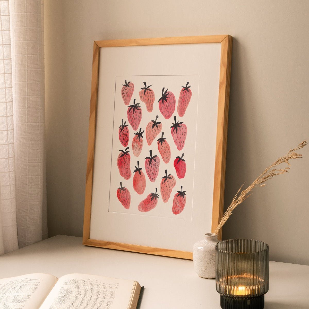 Framed original painting of pink and red strawberries, styled above a stack of books in a cozy home interior