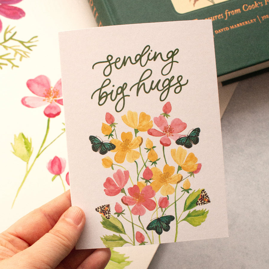 Hand holding a card with floral design and text 'Sending big hugs' on a surface with books in the background.