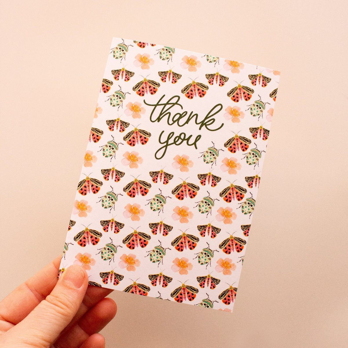 Hand holding a 'thank you' card with bug pattern on a beige background