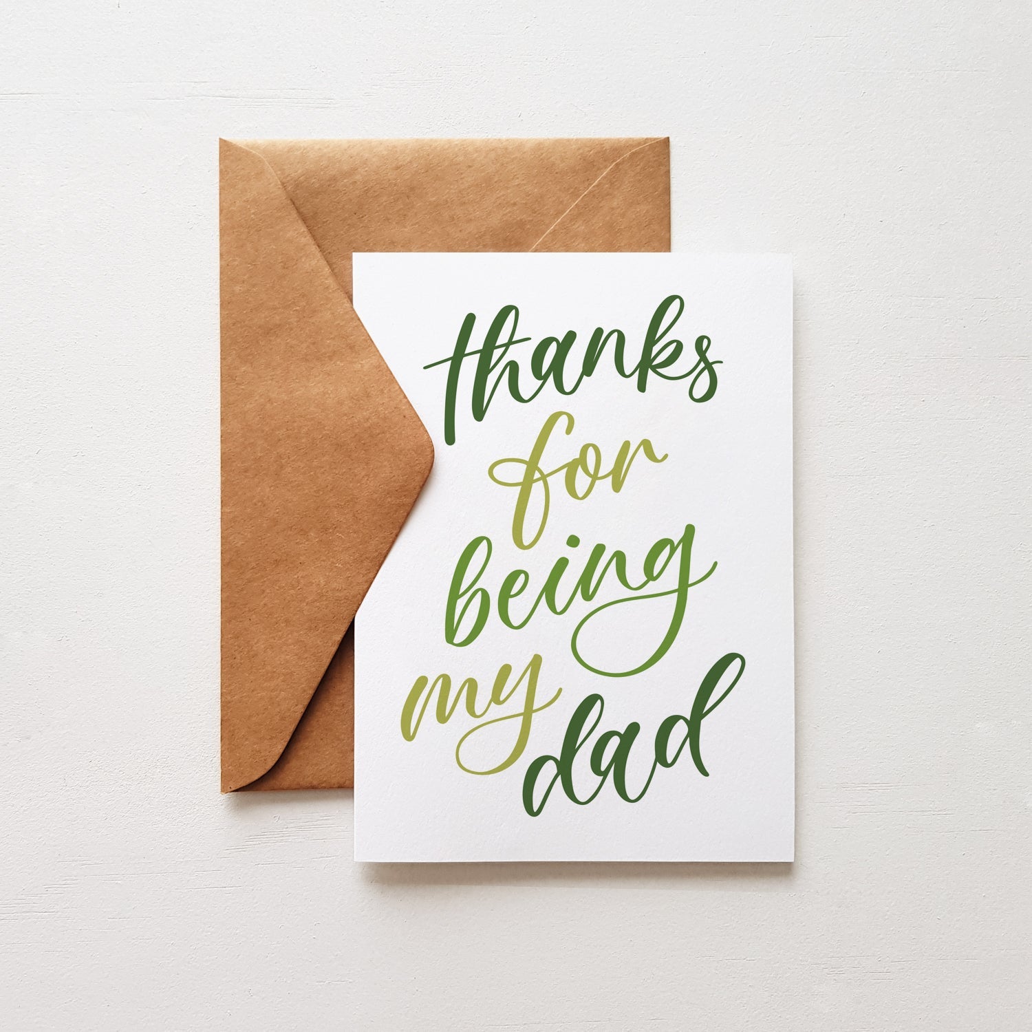 Green text 'Thanks for being my dad' on a white greeting card with a brown envelope