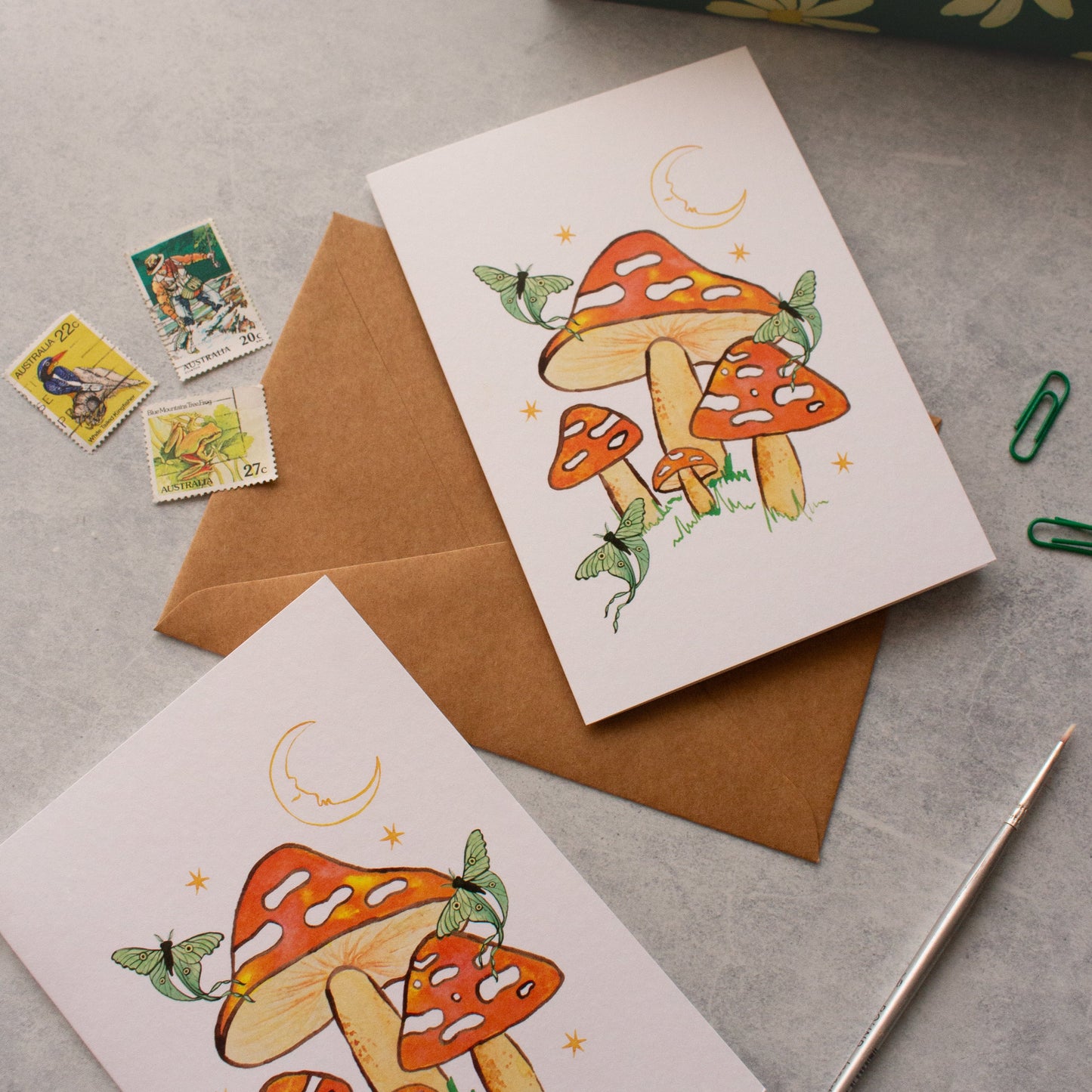 Two greeting cards with mushroom illustrations on a grey surface with stamps and an envelope.