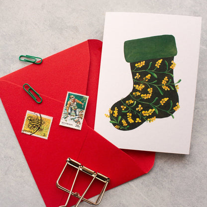 Christmas card with green stocking design, red envelope, and gold clips on grey surface