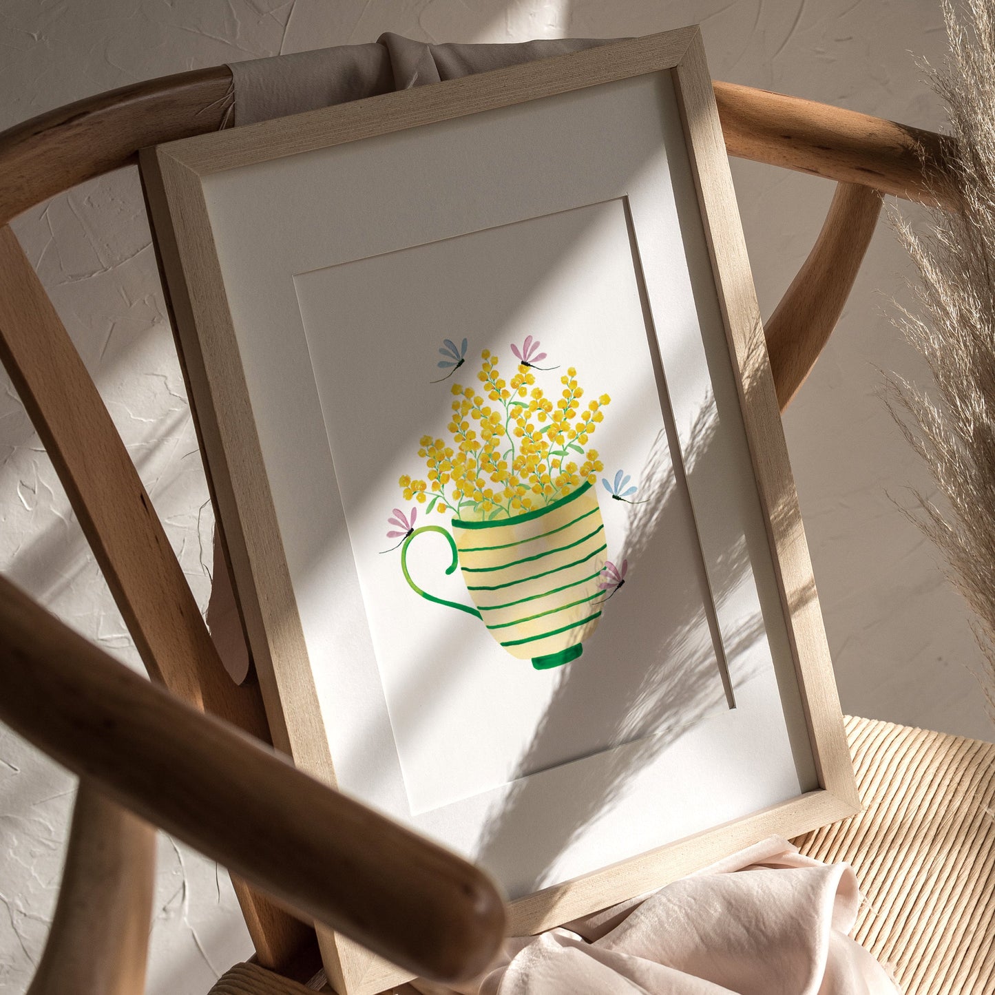 A framed artwork featuring a green striped tea cup filled with yellow wattle flowers, light purple and blue dragonflies, on a white background.