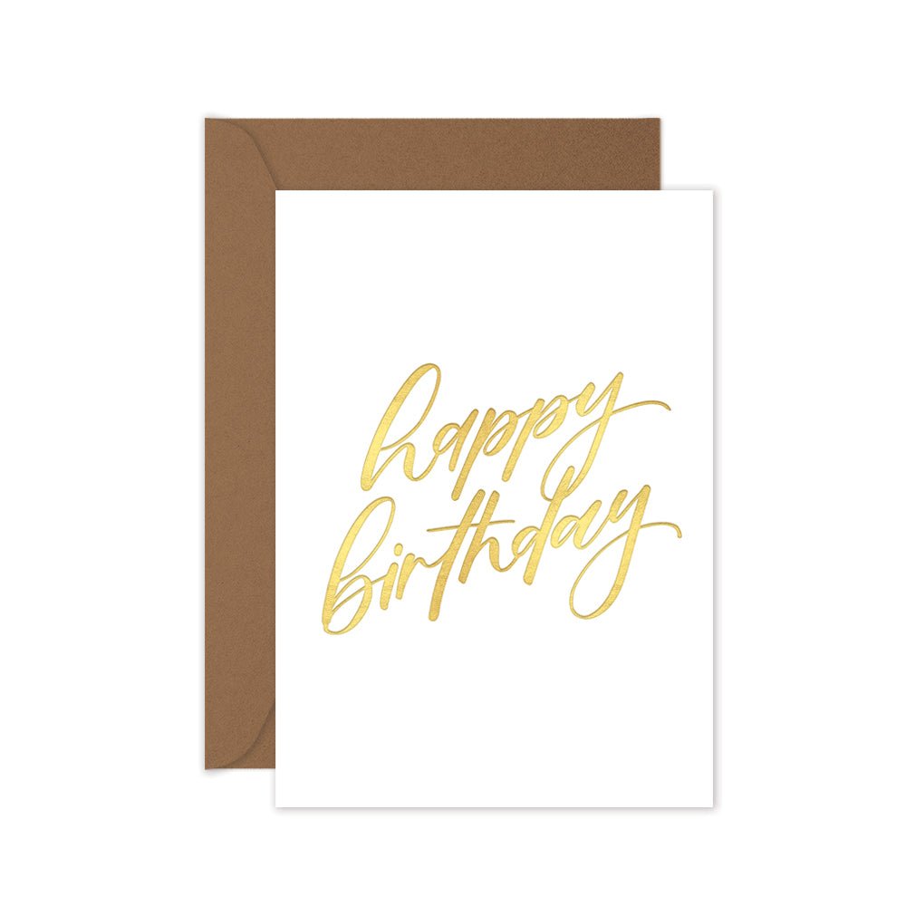 A folded white birthday card with 'Happy Birthday' hand lettering in gold, accompanied by a brown kraft envelope.