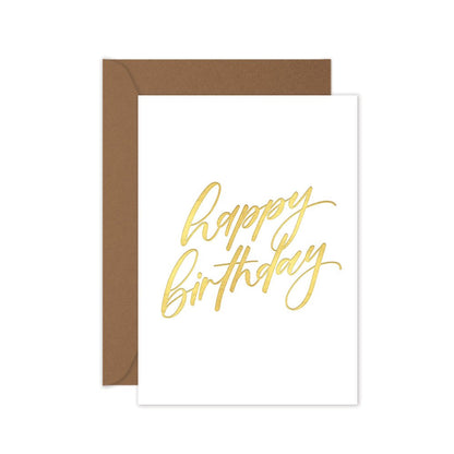 A folded white birthday card with 'Happy Birthday' hand lettering in gold, accompanied by a brown kraft envelope.