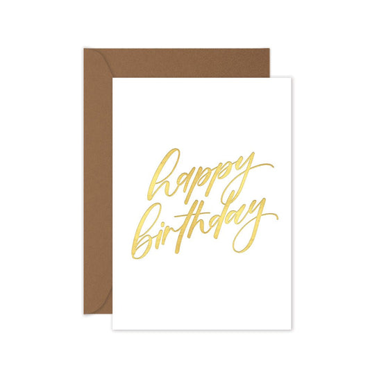 A folded white birthday card with 'Happy Birthday' hand lettering in gold, accompanied by a brown kraft envelope.