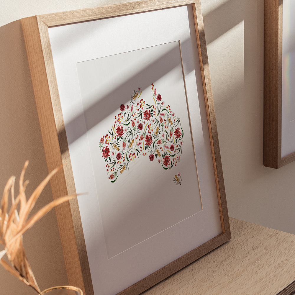 A framed art print of the Australian map, decorated with a floral pattern in red, greens, and yellow, placed on a wooden surface.